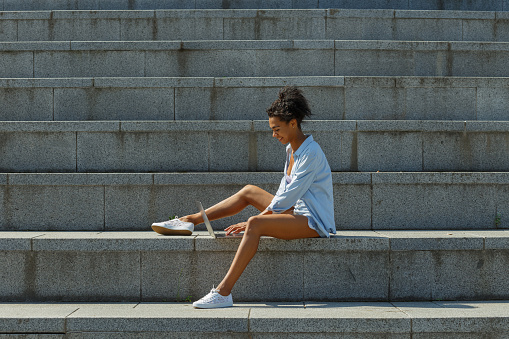 Positive mixed race young woman student in stylish clothes types on modern laptop sitting on large stone stairs on sunny summer day