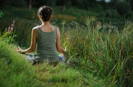 Woman doing meditation and relax at blooming spring lush meadow with green grass. back view