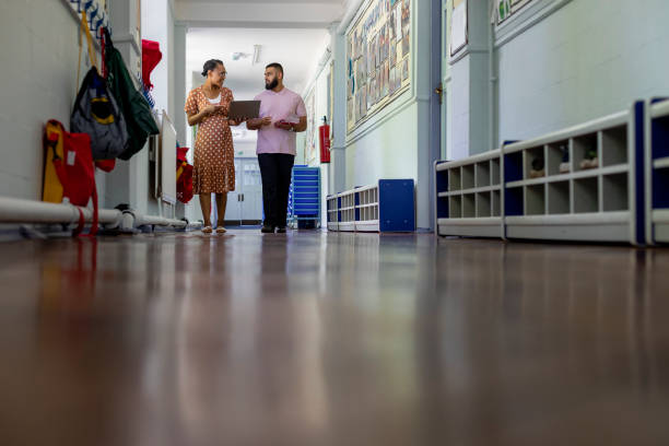 Sharing their Expertise Primary school teachers looking over lesson materials together in the North East of England while walking down a hallway. primary school assembly stock pictures, royalty-free photos & images