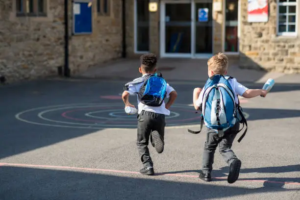 Rear view of two boys running in their school yard in the North East of England. They are running towards the door with their backpacks on.
