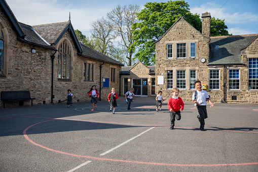 Rear view of children running in their school yard in the North East of England. They are all running away from the door with their backpacks on.