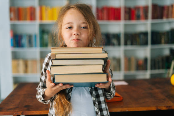 ritratto medio di una bella bambina delle elementari che tiene una pila di libri in biblioteca a scuola guardando la macchina fotografica. - elementary age focus on foreground indoors studio shot foto e immagini stock