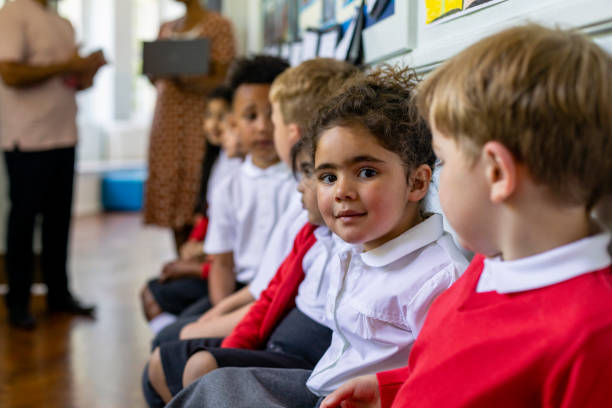 Students Waiting Together Primary school students sitting together in a line, one of the girls is looking at the camera smiling while at school in the North East of England. northeastern england stock pictures, royalty-free photos & images