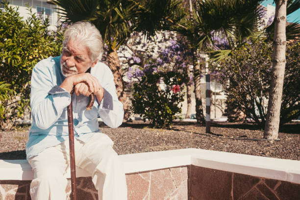 istock Old senior man with hands over the cane sitting in public park with sad expression. Elderly retiree with back pain using walking stick. Tree and flowers on background 1346505979