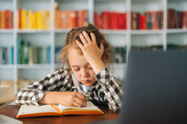 istock Close-up front view of exhausted cute elementary child school girl doing writing homework holding pen sitting at table with laptop and paper notebook 1346506924