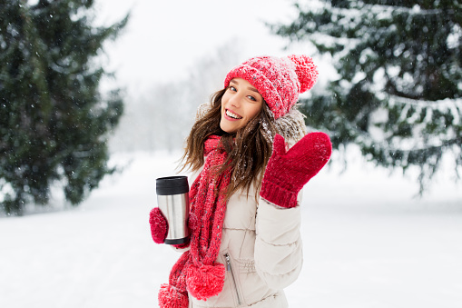 people, season, drinks and christmas concept - happy teenage girl or young woman with hot drink in tumbler outdoors in winter park and waving hand