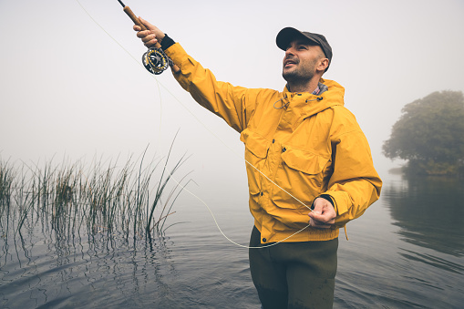 Male wearing cap and a yellow fishing jacket while fly-fishing on a early and misty morning. The fog is just about to evaporate as the warming sun rays break through to warm up the beautiful Furesø, the deepest lake in Denmark. .