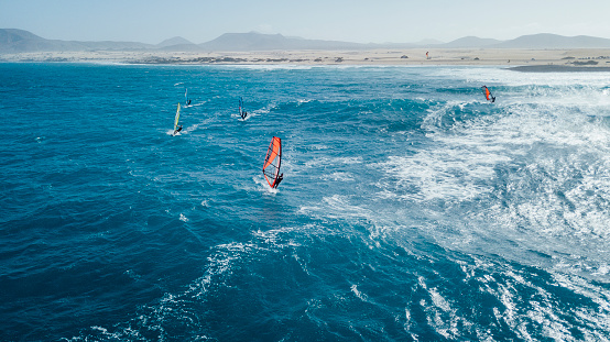 A drone captures wind surfers catching waves along the coastal line of Fuerteventura, big blue open waters with high winds.