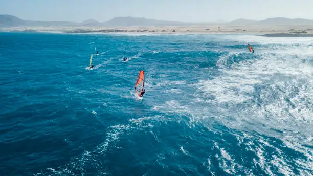 A drone captures wind surfers catching waves along the coastal line of Fuerteventura, big blue open waters with high winds.