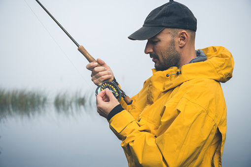 Male wearing cap and a yellow fishing jacket while fly-fishing on a early and misty morning. The fisherman is focused on choosing the right fly for the current situation. The fog is just about to evaporate as the warming sun rays break through to warm up the beautiful Furesø, the deepest lake in Denmark. .