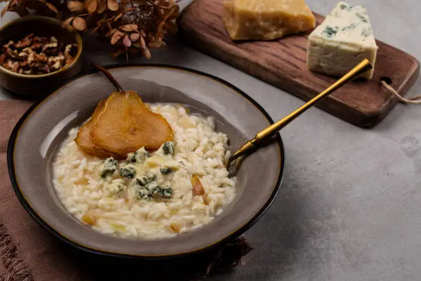 Pear and gorgonzola risotto with walnuts on grey table. Hard and soft cheeses on background. Copy space.