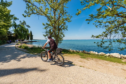 Lazise, Italy - May 26, 2021: Coast of Lake Garda (Lago di Garda) with a beach and bicycle and pedestrian lane that connects the towns of Lazise, Cisano, Bardolino and Garda. Verona province, Veneto, Italy, Europe. A group of tourists and locals walking or cycling on a sunny spring day.