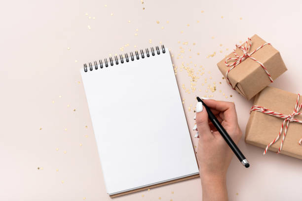 istock Female hand write hold notebook blank paper mockup, golden stars confetti, gift boxes on beige background. 1346517639