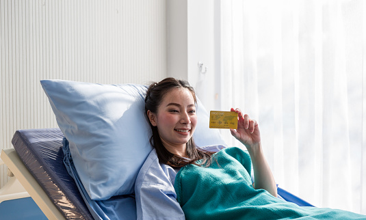 Asian female patient show credit card in hand while lying on bed. Woman patient prepare pay medical expenses. Female patient with credit or health card for medical treatment. Health Insurance concept