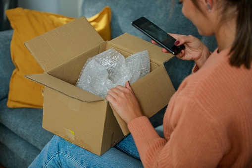 High angle view of young woman using smart phone and taking a photo of a package that she opened, as a proof of delivery confirmation.