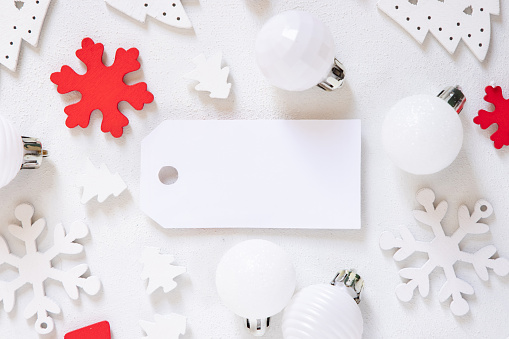 Blank gift tag with white and red Christmas decorations top view on white table. Winter composition with blank label card Mockup, copy space
