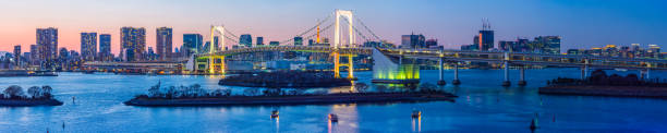 Tokyo Rainbow Bridge over bay skyscrapers illuminated sunset panorama Japan The soaring span of the Rainbow Bridge crossing over Tokyo Bay from Odaiba past the iconic spire of the Tokyo Tower towards downtown skyscrapers illuminated against sunset skies, Japan. tokyo prefecture tokyo tower japan night stock pictures, royalty-free photos & images