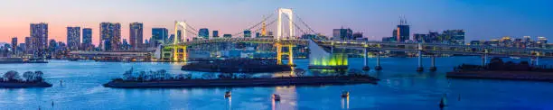 The soaring span of the Rainbow Bridge crossing over Tokyo Bay from Odaiba past the iconic spire of the Tokyo Tower towards downtown skyscrapers illuminated against sunset skies, Japan.