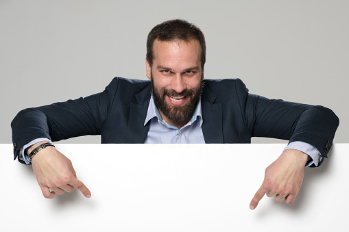 Portrait of smiling mature businessman pointing downwards while leaning on white polystyrene board against gray background.