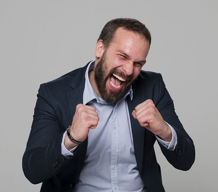 Portrait of excited mature businessman clenching fist while standing against gray background.