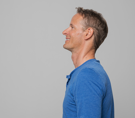 Side view of smiling mature man in blue t-shirt looking up while standing against gray background.