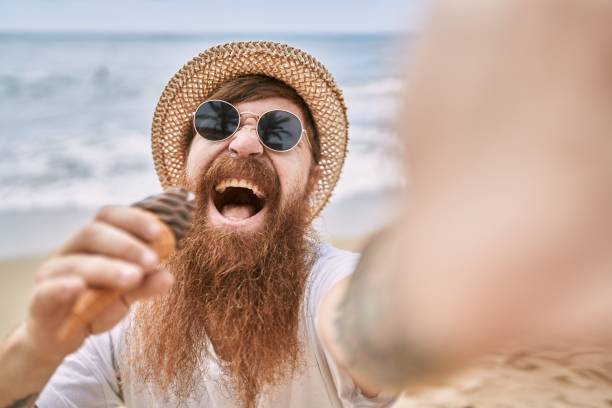 istock Young redhead man eating ice cream make selfie by the camera at the beach. 1346535685
