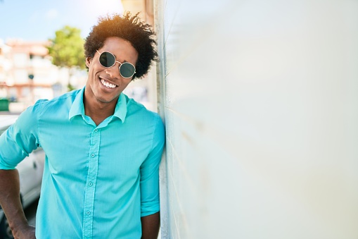 Young handsome african american man wearing casual clothes and sunglasses smiling happy. Leaning on the wall with smile on face at town street.