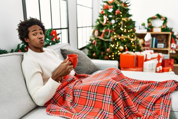 istock Young african american man sitting on the sofa drinking coffee by christmas tree looking at the camera blowing a kiss on air being lovely and sexy. love expression. 1346536947