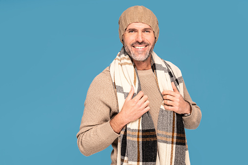 Fashionable bearded handsome man wearing big warm scarf and winter cap, smiling to the camera. Blue pastel studio background. A lot of copy space.