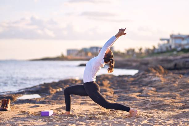 young beautiful sportwoman practicing yoga. coach teaching warrior pose at the beach - stretch beach imagens e fotografias de stock