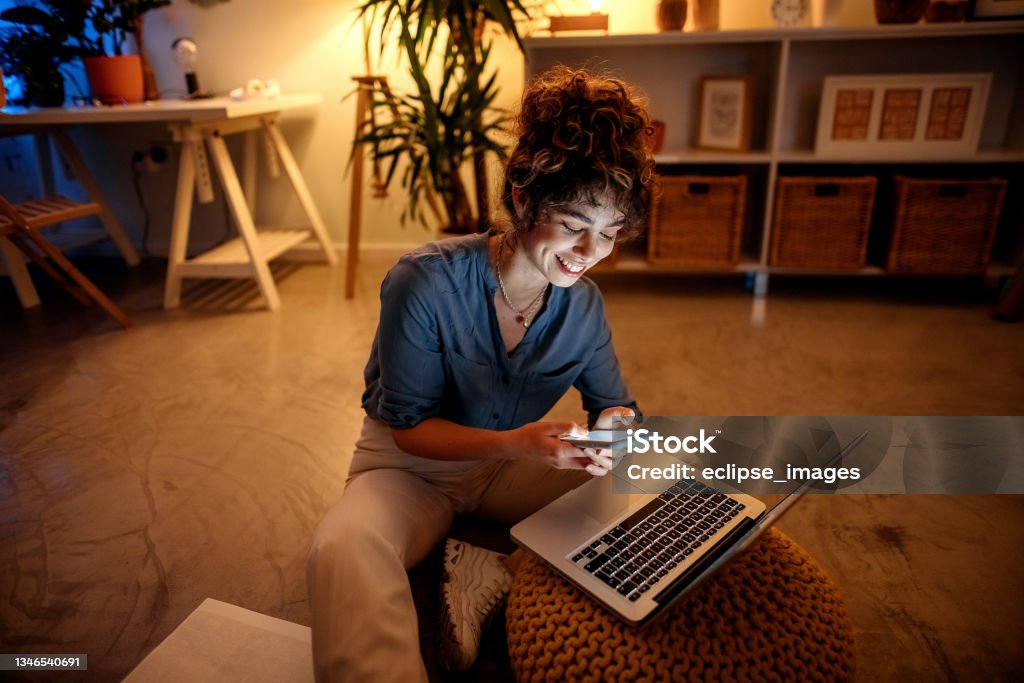 Young businesswoman working from home at night Young beautiful woman sits on the floor in the living room and uses a laptop and a mobile phone People Stock Photo