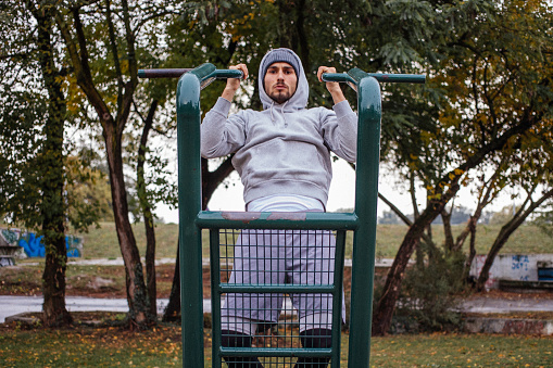 A young men in a gray tracksuit on an autumn rainy day exercising on a gymnastic device in a public park surrounded by an urban environment