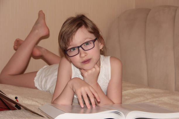 A child with glasses is lying on the bed and reading a book. Distance learning.