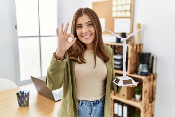 Photo of Young brunette woman holding solar windmill for renewable electricity at the office doing ok sign with fingers, smiling friendly gesturing excellent symbol