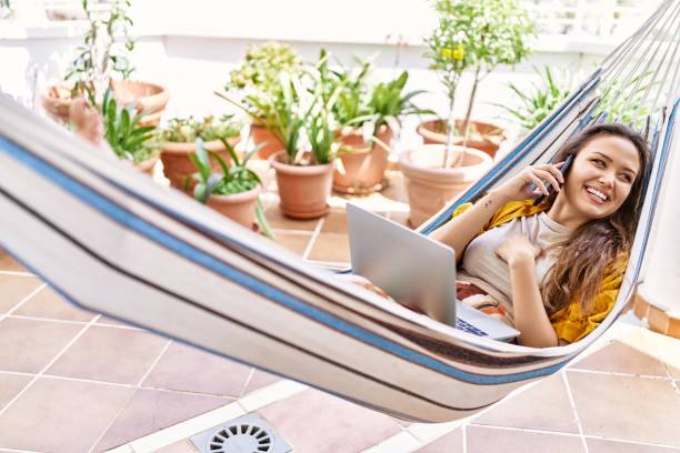 jeune fille hispanique parlant sur le smartphone et utilisant un ordinateur portable allongé sur un hamac à la terrasse. - hamac telephone homme photos et images de collection