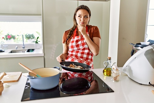 Beautiful young brunette woman wearing apron cooking at the kitchen serious face thinking about question with hand on chin, thoughtful about confusing idea