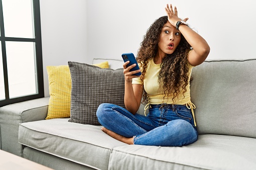 Beautiful hispanic woman sitting on the sofa at home using smartphone surprised with hand on head for mistake, remember error. forgot, bad memory concept.