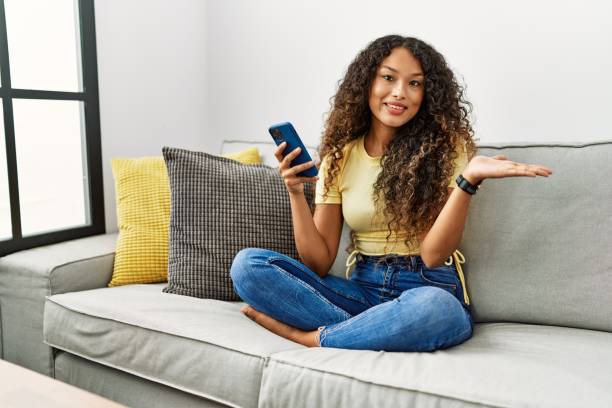 istock Beautiful hispanic woman sitting on the sofa at home using smartphone smiling cheerful presenting and pointing with palm of hand looking at the camera. 1346549963