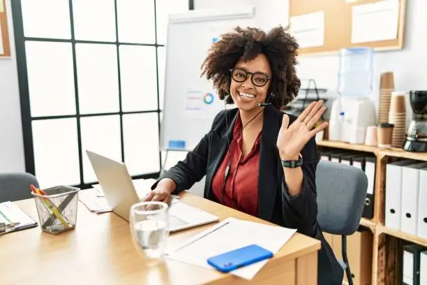Photo of African american woman with afro hair working at the office wearing operator headset waiving saying hello happy and smiling, friendly welcome gesture