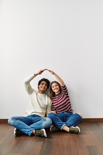 Young hispanic couple doing roof symbol with hands sitting on the floor at empty new home.