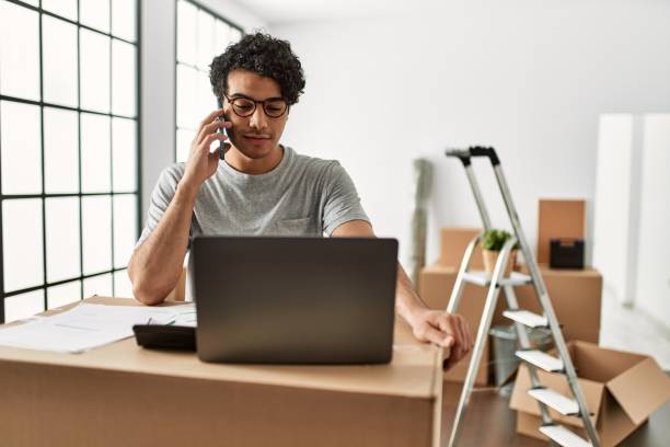 Young hispanic man talking on the smartphone using laptop at new home. Young hispanic man talking on the smartphone using laptop at new home. company relocation stock pictures, royalty-free photos & images