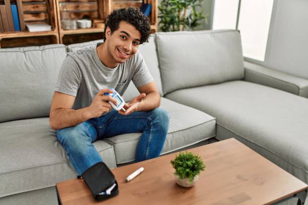 Young hispanic diabetic man measuring glucose sitting on the sofa at home. stock photo