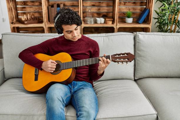 istock Young hispanic man playing classical guitar sitting on the sofa at home. 1346554054