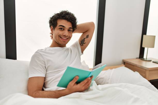 istock Young hispanic man reading book sitting on the bed at bedroom. 1346554116
