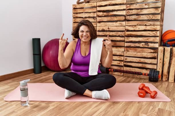 Middle age hispanic woman sitting on training mat at the gym shouting with crazy expression doing rock symbol with hands up. music star. heavy music concept.