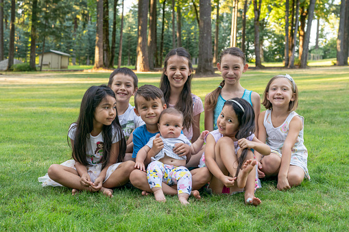 A multi-ethnic group of children of various ages sit in the grass and pose for a photograph while at a family reunion. The affectionate cousins are happy and smiling directly at the camera. Aging process, development, diversity, family and friendship concepts.