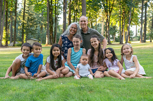 A mixed race senior couple embrace while posing with their grandchildren outdoors at a family reunion. The happy retired husband and wife are filled with gratitude and excitement as they enter retirement. Their happy grandchildren vary in ages and ethnicities.