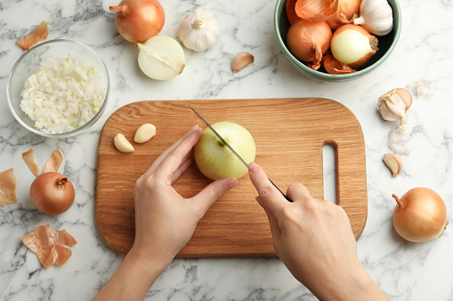 Woman cutting fresh onion on wooden board at white marble table, top view