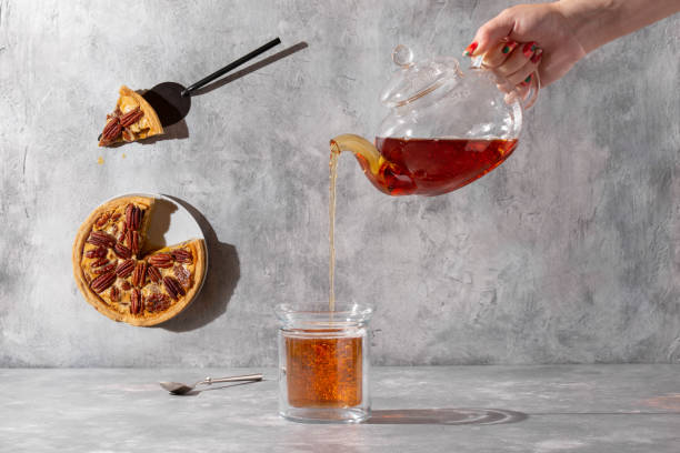 fête du thé à la tarte aux noix de pécan. coup créatif de verser du thé. femme versant à la main du théier en verre dans une tasse sur fond gris béton. - five oclock tea photos et images de collection