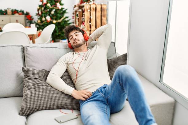 istock Young hispanic man celebrating christmas using headphones lying on the sofa at home. 1346561477